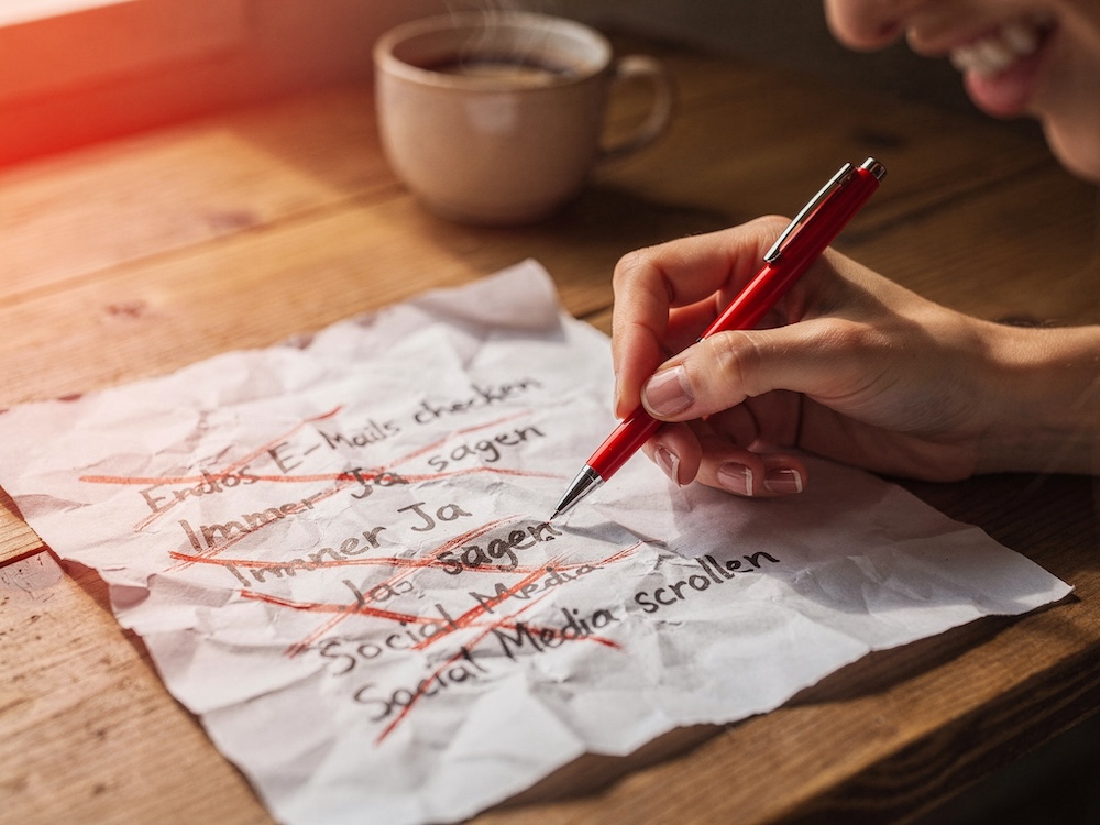 "Person streicht mit rotem Stift Einträge von einer Liste durch, Gesichtsausdruck wirkt befreit und erleichtert - symbolisiert die Kraft der Streichliste, Ballast loszulassen statt immer mehr hinzuzufügen"